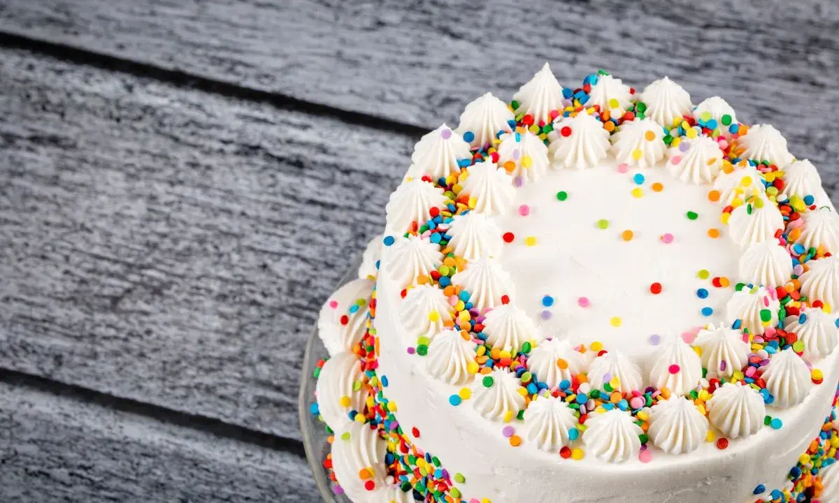 Torta compleanno bianca con glassa, decorata con ciuffi di panna e confettini colorati.