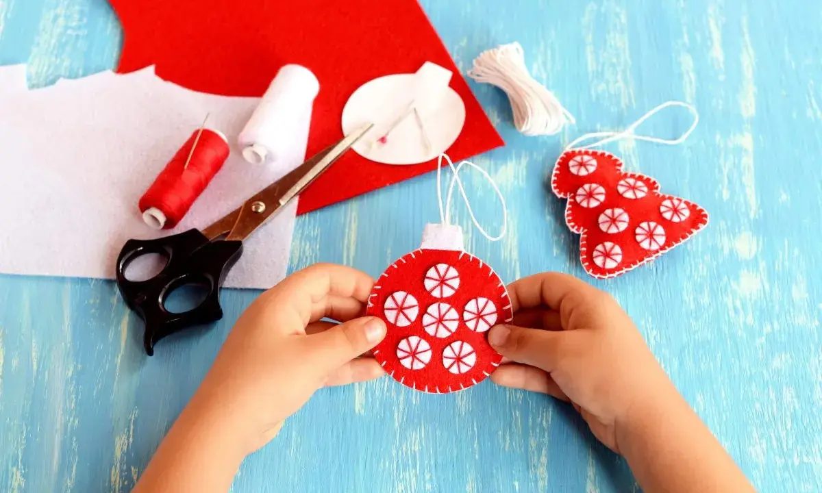 Bambino crea decorazioni natalizie in feltro. Materiali per attività natalizie per bambini: forbici, filo rosso e bianco, feltro rosso e bianco.