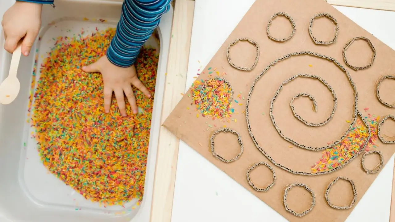 Bambino esplora riso colorato in vaschetta, parte di giochi sensoriali tattili. Cartone con spirale e cerchi decorati con riso.