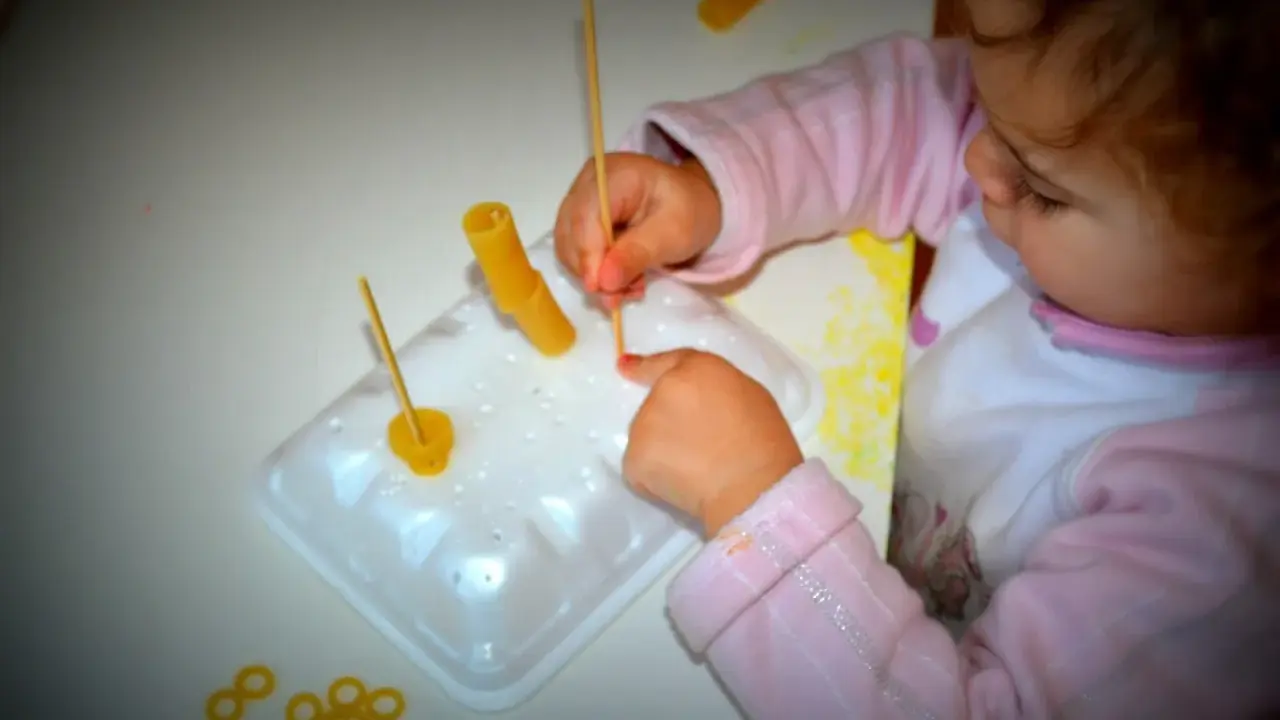 Bambina di 3 anni si diverte con giochi creativi: infila pasta su stecchini, sviluppando manualità e fantasia.
