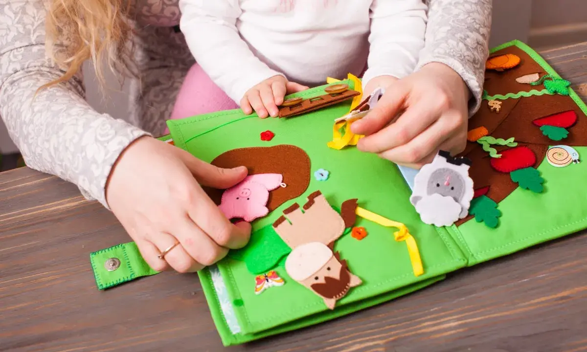 Mani di adulto e bambino esplorano un colorato libro sensoriale in feltro fai da te, con animali e elementi naturali.