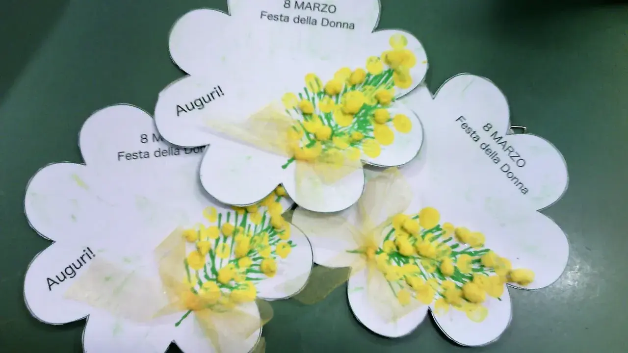Lavoretto festa della donna infanzia: tre fiori di carta con mimose dipinte e fiocchi gialli. Scritte "Auguri!" e "8 Marzo Festa della Donna".