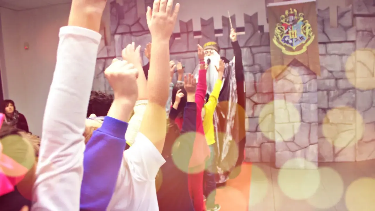 Bambini entusiasti alzano le mani per imparare trucchi di magia facili, con un mago sullo sfondo e uno stemma di Hogwarts.