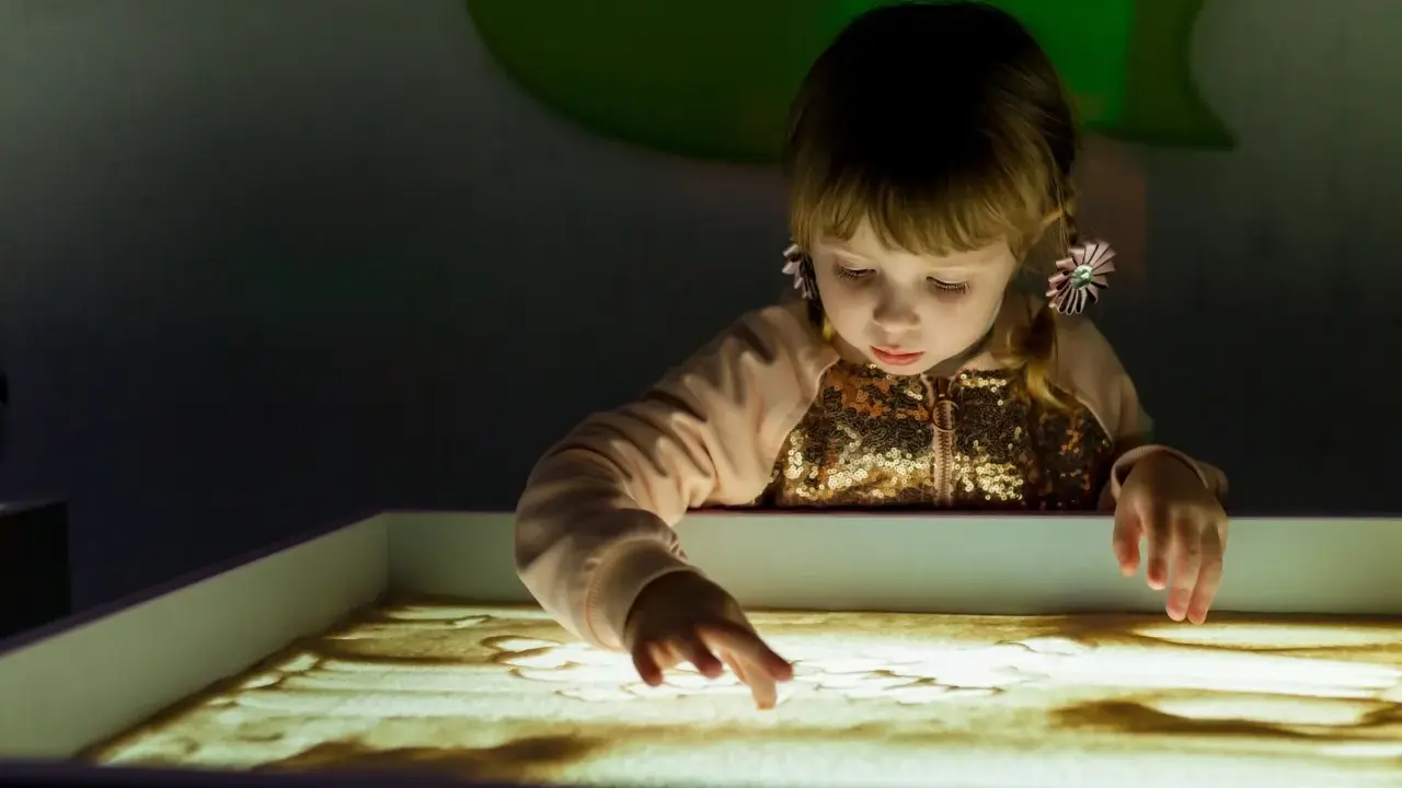 Bambina con i capelli biondi e fiocchi rosa crea disegni nella sabbia illuminata, un momento di sandplay therapy.