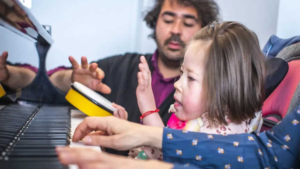 Bambina e terapista suonano il piano, con un tamburello giallo. Esempi di musicoterapia che promuovono l'interazione e l'espressione.