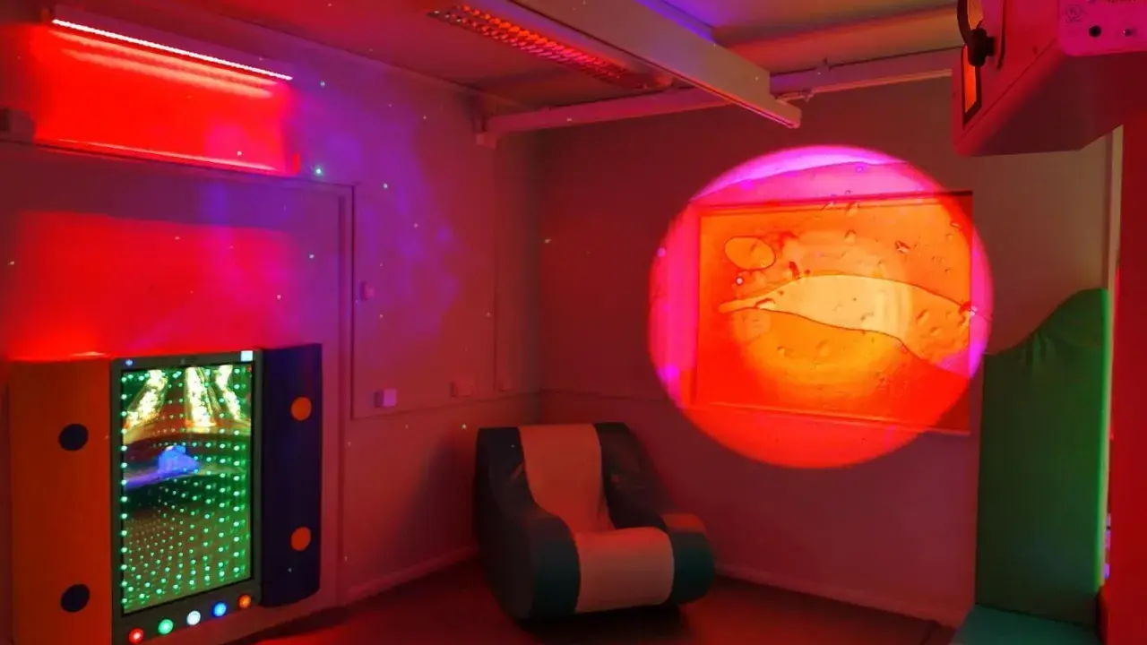 Una stanza snoezelen con luci soffuse, un pannello interattivo luminoso, una poltrona comoda e un proiettore di immagini rilassanti.