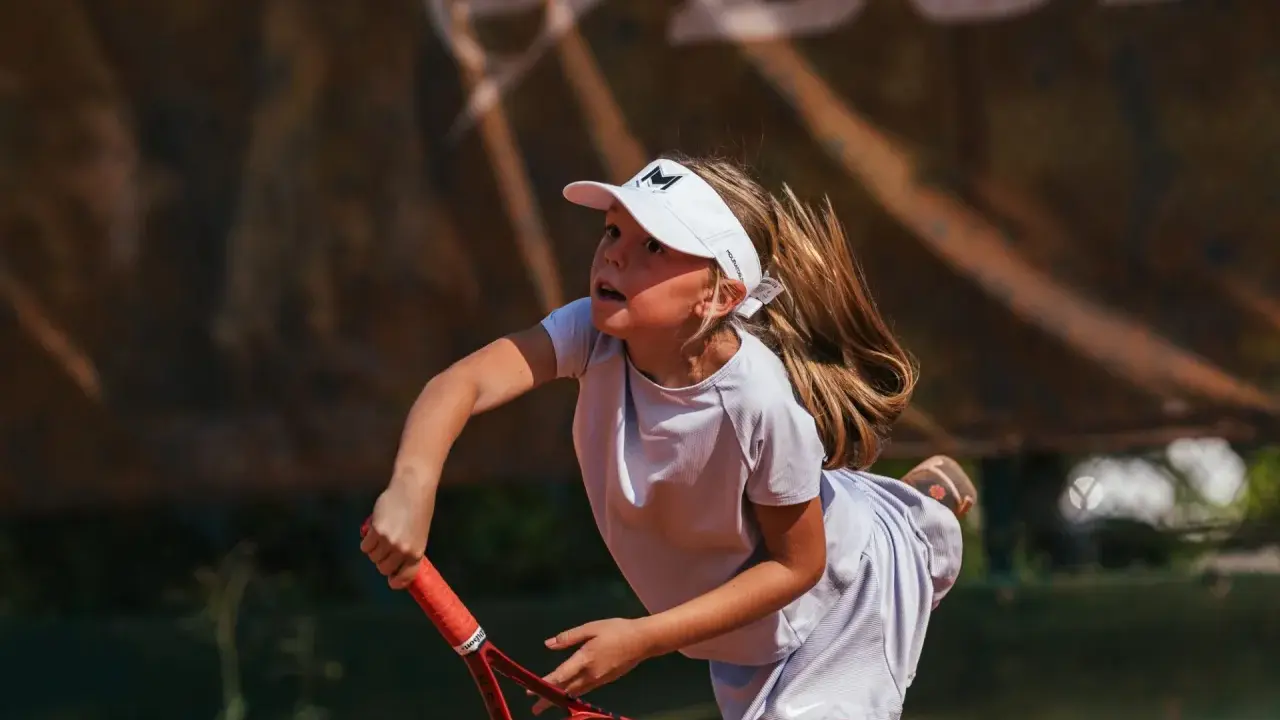 Bambina concentrata durante un servizio di tennis, con la racchetta pronta a colpire la palla.