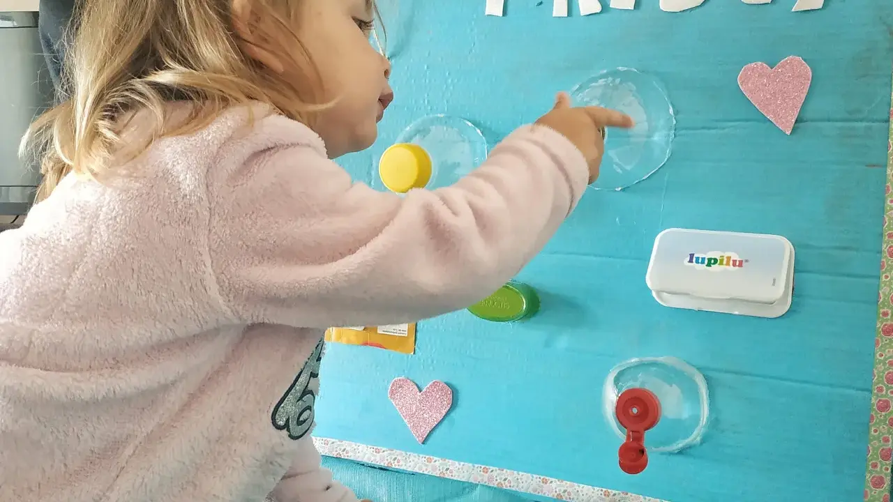Bambina gioca con giochi motricità fine fai da te: un pannello azzurro con lettere glitterate, cuori rosa e oggetti da manipolare.