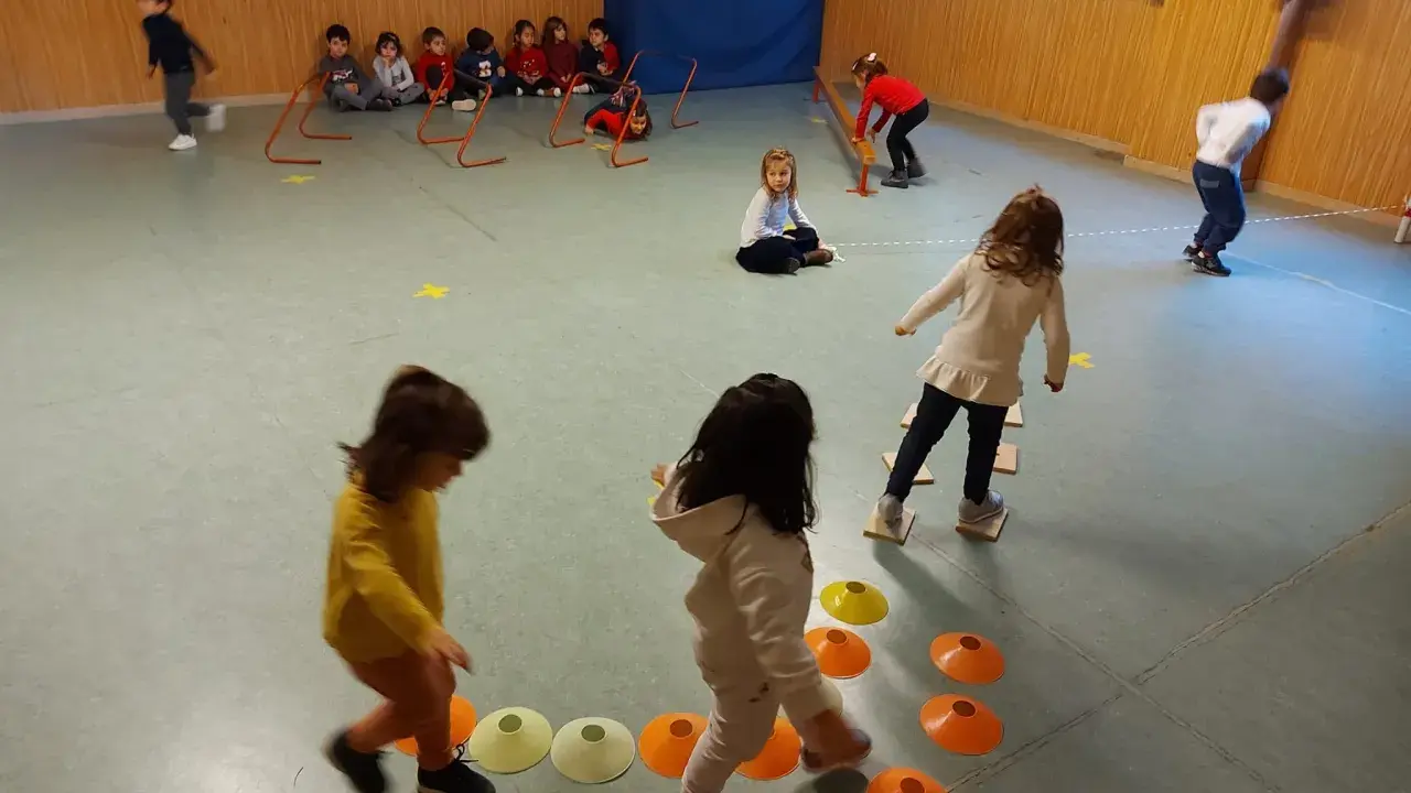 Bambini si divertono con giochi psicomotricità 3 anni: corrono, saltano ostacoli e camminano su coni colorati in palestra.