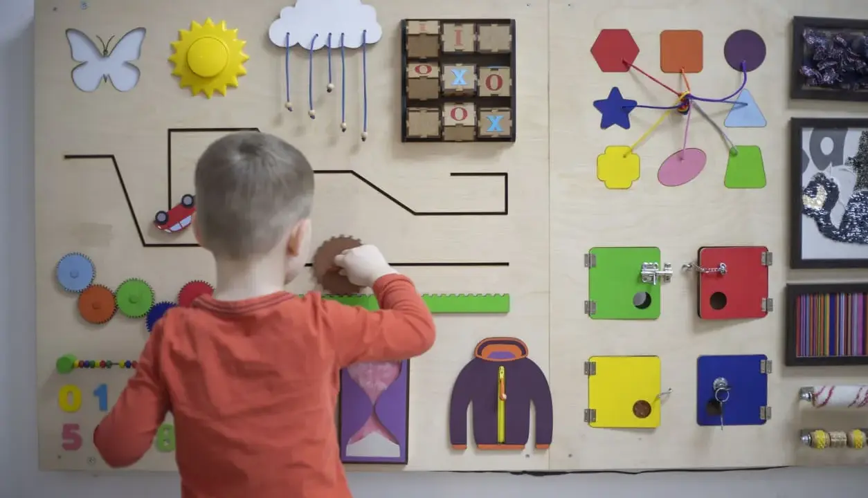 Bambino gioca con un pannello sensoriale fai da te: ingranaggi, lucchetti, forme e un gioco del tris.