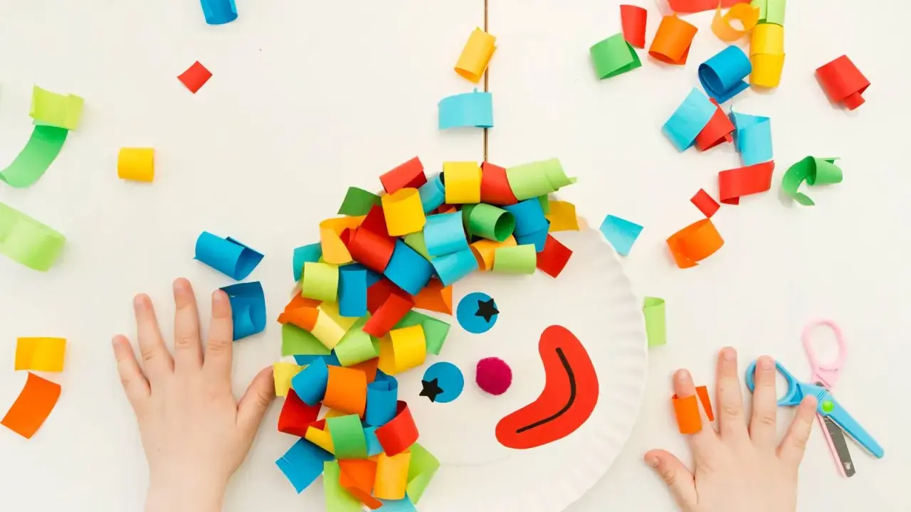 Bambini creano un clown con carta colorata, un'idea per lavoretti di Carnevale. Mani piccole e forbici rosa completano la scena.