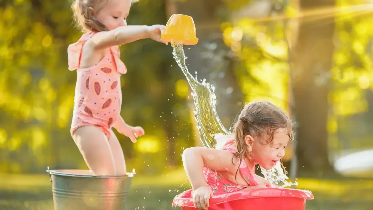 Bambine si divertono con giochi d'acqua al nido, una spruzza l'altra con un secchiello giallo.