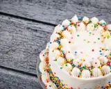 Torta compleanno bianca con glassa, decorata con ciuffi di panna e confettini colorati.