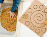 Bambino esplora riso colorato in vaschetta, parte di giochi sensoriali tattili. Cartone con spirale e cerchi decorati con riso.