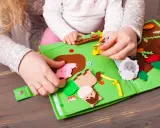 Mani di adulto e bambino esplorano un colorato libro sensoriale in feltro fai da te, con animali e elementi naturali.