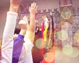 Bambini entusiasti alzano le mani per imparare trucchi di magia facili, con un mago sullo sfondo e uno stemma di Hogwarts.