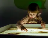 Bambina con i capelli biondi e fiocchi rosa crea disegni nella sabbia illuminata, un momento di sandplay therapy.