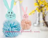Coniglietti con pompon di lana e teste di coniglio origami. Uno è azzurro, l'altro rosa, con fiori gialli sullo sfondo.