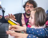Bambina e terapista suonano il piano, con un tamburello giallo. Esempi di musicoterapia che promuovono l'interazione e l'espressione.