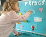 Bambina gioca con giochi motricità fine fai da te: un pannello azzurro con lettere glitterate, cuori rosa e oggetti da manipolare.