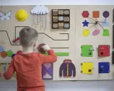 Bambino gioca con un pannello sensoriale fai da te: ingranaggi, lucchetti, forme e un gioco del tris.