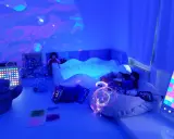 Stanza sensoriale autismo con piscina di palline, luci soffuse, pupazzi e pannelli interattivi per stimolare i sensi.