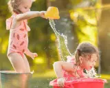 Bambine si divertono con giochi d'acqua al nido, una spruzza l'altra con un secchiello giallo.