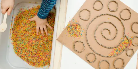 Bambino esplora riso colorato in vaschetta, parte di giochi sensoriali tattili. Cartone con spirale e cerchi decorati con riso.