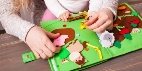 Mani di adulto e bambino esplorano un colorato libro sensoriale in feltro fai da te, con animali e elementi naturali.