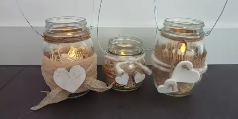 Tre vasetti lanterne fai da te decorati con cuori e spago, con candele accese all'interno per un'atmosfera calda.