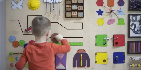 Bambino gioca con un pannello sensoriale fai da te: ingranaggi, lucchetti, forme e un gioco del tris.