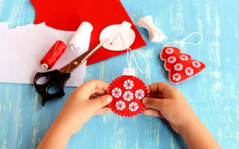 Bambino crea decorazioni natalizie in feltro. Materiali per attività natalizie per bambini: forbici, filo rosso e bianco, feltro rosso e bianco.