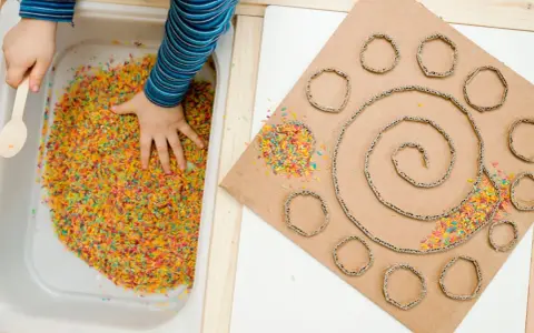 Bambino esplora riso colorato in vaschetta, parte di giochi sensoriali tattili. Cartone con spirale e cerchi decorati con riso.
