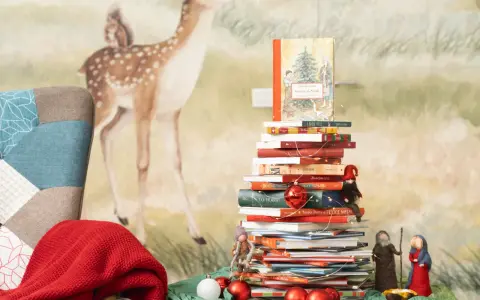Una pila di libri per le letture di Natale per bambini, decorata con lucine e palline rosse, con un folletto e statuine del presepe.
