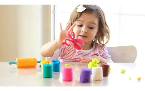 Bambina gioca con pasta modellabile colorata, forbici rosa e mattarello arancione. Giochi di manipolazione Montessori per sviluppare creatività.