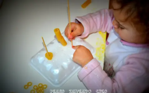 Bambina di 3 anni si diverte con giochi creativi: infila pasta su stecchini, sviluppando manualità e fantasia.