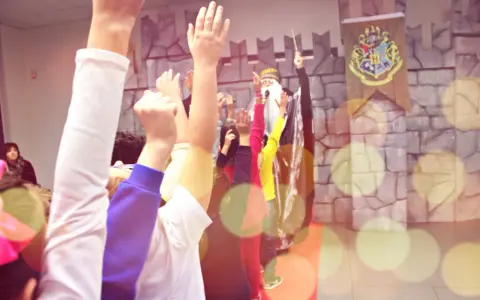Bambini entusiasti alzano le mani per imparare trucchi di magia facili, con un mago sullo sfondo e uno stemma di Hogwarts.