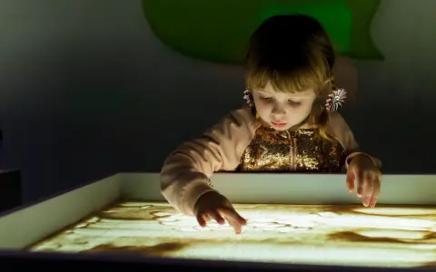 Bambina con i capelli biondi e fiocchi rosa crea disegni nella sabbia illuminata, un momento di sandplay therapy.