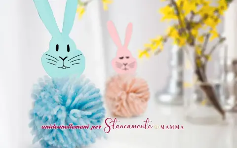 Coniglietti con pompon di lana e teste di coniglio origami. Uno è azzurro, l'altro rosa, con fiori gialli sullo sfondo.