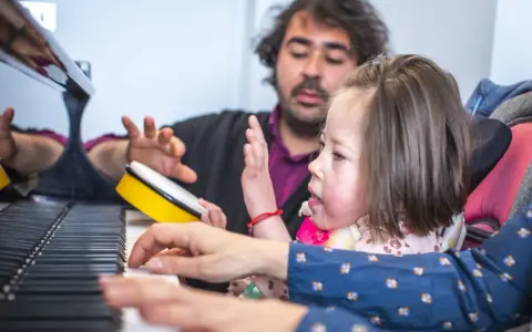Bambina e terapista suonano il piano, con un tamburello giallo. Esempi di musicoterapia che promuovono l'interazione e l'espressione.