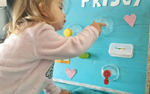 Bambina gioca con giochi motricità fine fai da te: un pannello azzurro con lettere glitterate, cuori rosa e oggetti da manipolare.