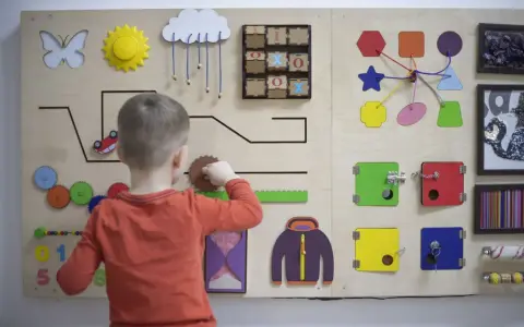 Bambino gioca con un pannello sensoriale fai da te: ingranaggi, lucchetti, forme e un gioco del tris.