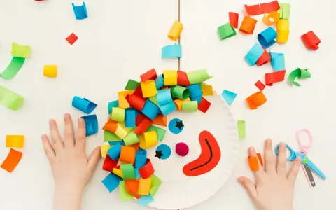 Bambini creano un clown con carta colorata, un'idea per lavoretti di Carnevale. Mani piccole e forbici rosa completano la scena.