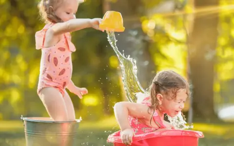 Bambine si divertono con giochi d'acqua al nido, una spruzza l'altra con un secchiello giallo.