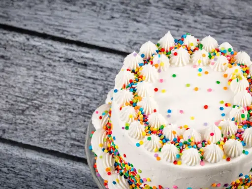 Torta compleanno bianca con glassa, decorata con ciuffi di panna e confettini colorati.