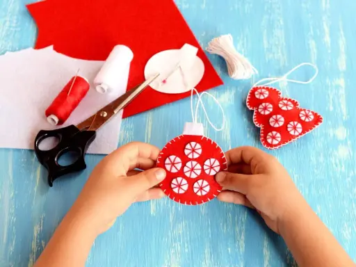 Bambino crea decorazioni natalizie in feltro. Materiali per attività natalizie per bambini: forbici, filo rosso e bianco, feltro rosso e bianco.