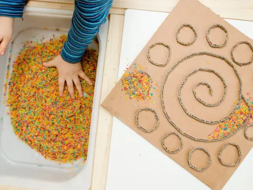 Bambino esplora riso colorato in vaschetta, parte di giochi sensoriali tattili. Cartone con spirale e cerchi decorati con riso.
