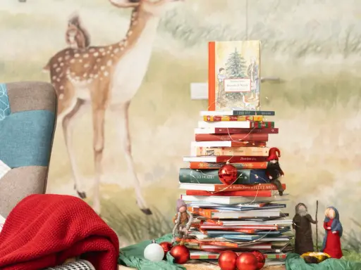 Una pila di libri per le letture di Natale per bambini, decorata con lucine e palline rosse, con un folletto e statuine del presepe.