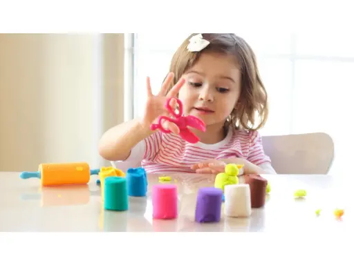 Bambina gioca con pasta modellabile colorata, forbici rosa e mattarello arancione. Giochi di manipolazione Montessori per sviluppare creatività.