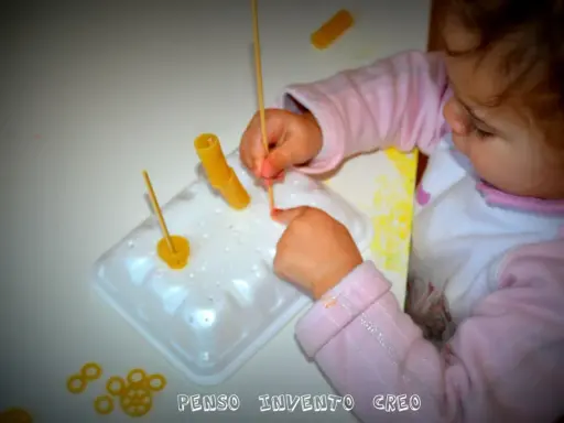 Bambina di 3 anni si diverte con giochi creativi: infila pasta su stecchini, sviluppando manualità e fantasia.